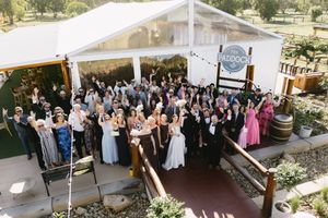 Wedding guests gather outdoors at a rustic vineyard venue with white chairs and wine barrels for the ceremony.