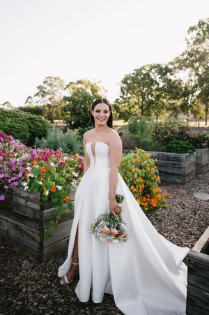 Bride in strapless gown poses in garden with colorful flowers and wooden planter boxes.