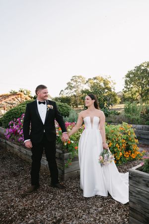 Newlyweds stand together in a garden with raised flower beds and vibrant orange blooms.
