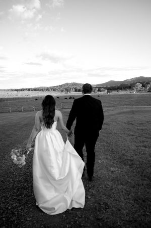 Black and white photo of newlyweds walking across open field with mountains in background.