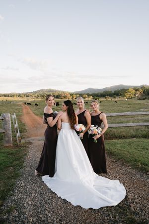 Bridal party in black dresses and white wedding gown posing on a gravel path with mountains in background.