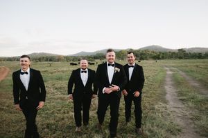 Groomsmen in black tuxedos standing in open field with mountain landscape.