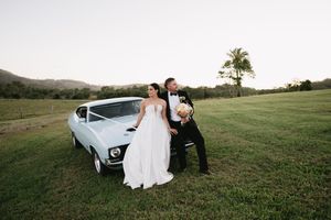 Wedding couple poses with vintage white car in scenic countryside setting.