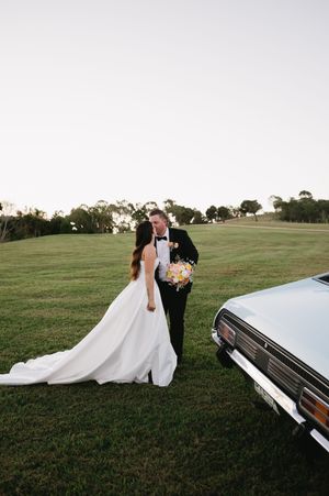 Romantic wedding couple poses near vintage car in grassy field at sunset.