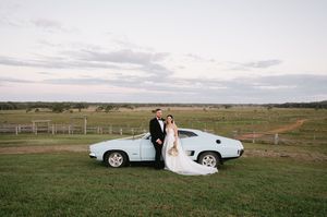 Wedding dress train trailing behind a white vintage car in an open field landscape.