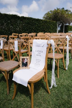 Wooden cross-back chairs with white ribbons and ceremony programs arranged on a grassy lawn for an outdoor wedding.