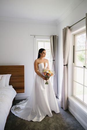 Bride in a white wedding dress stands near a window in an elegant room with gray curtains and dark carpet.