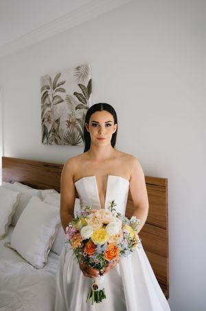 Elegant white wedding dress with plunging neckline and colorful spring bouquet in a modern bedroom setting.