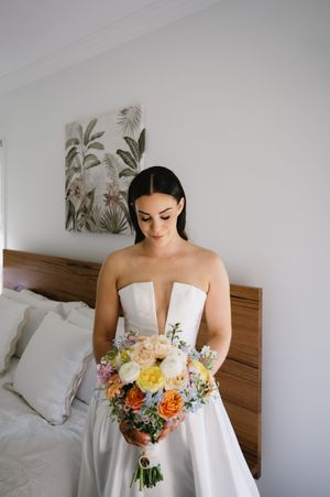 Bride in white strapless gown holding colorful peach and yellow bouquet in bedroom setting.