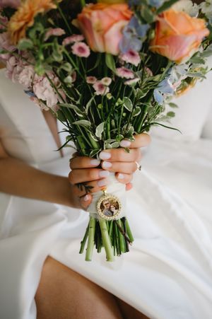 Close-up details of pastel wedding bouquet with peach roses and greenery against white fabric.