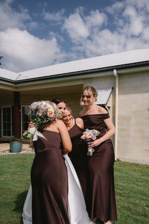 Bridesmaids in chocolate brown dresses share laughs on a sunny lawn outside a white building.