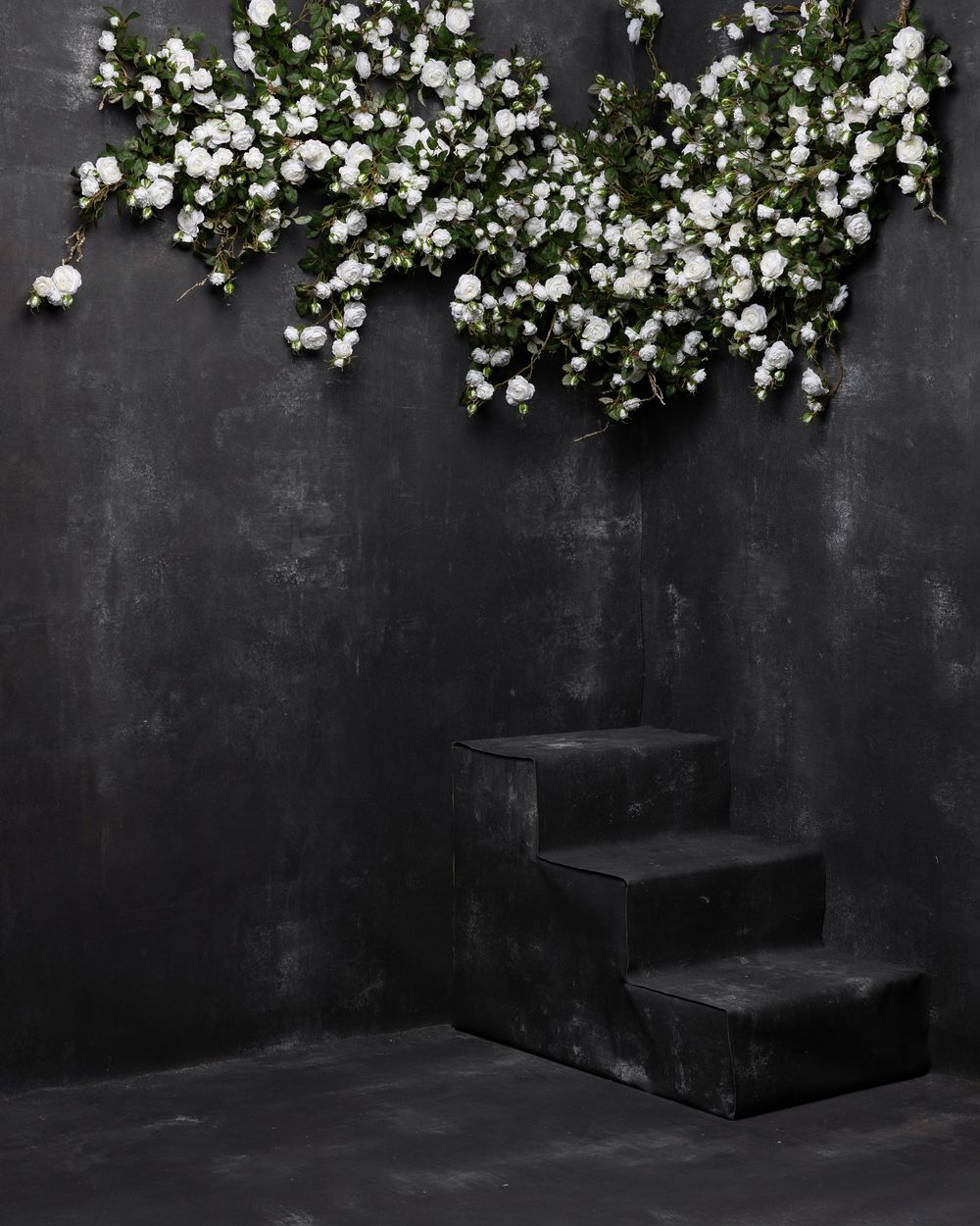 White flowers cascade over a dark wall above black wooden steps in a moody, minimalist setting.
