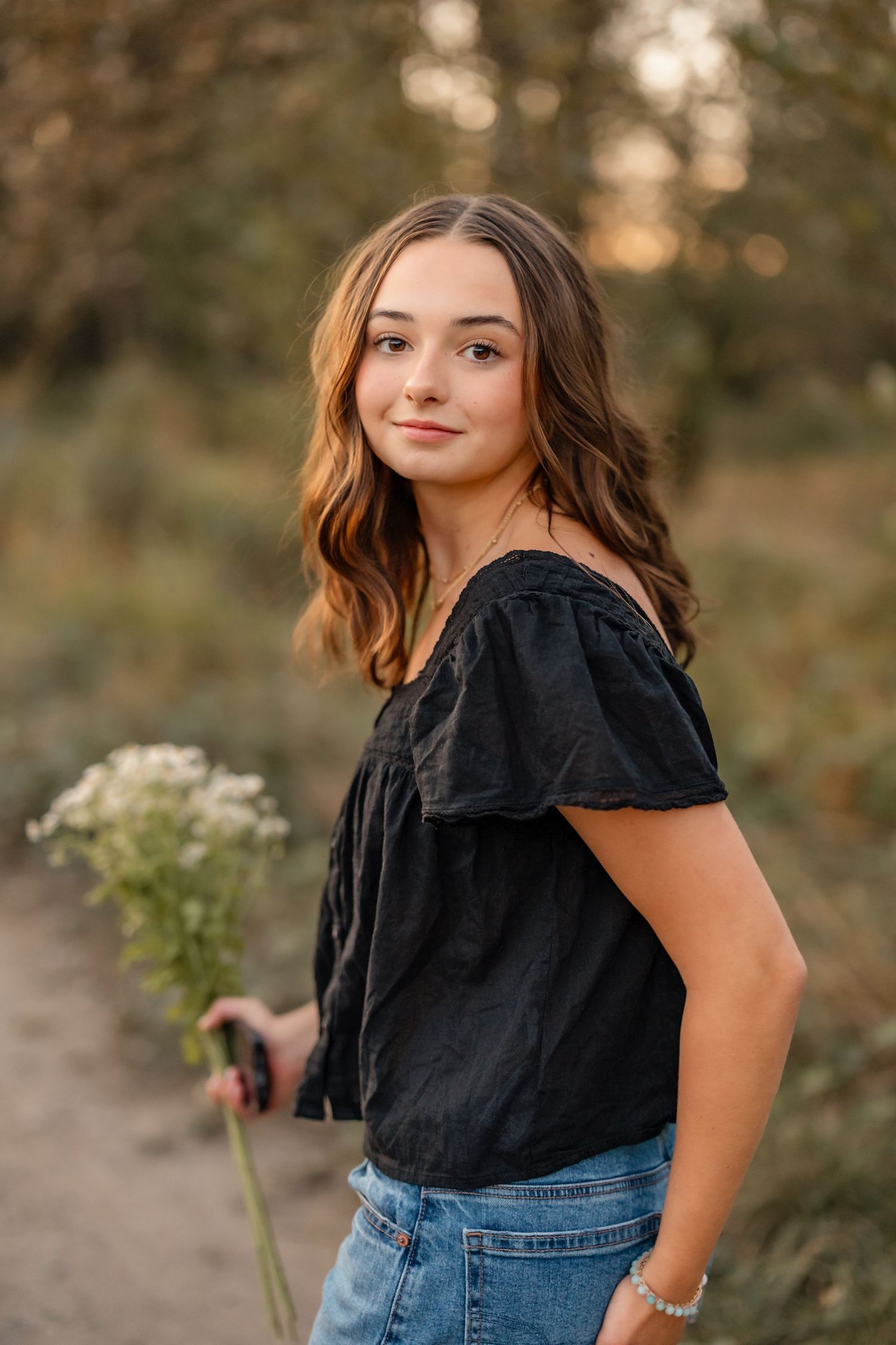 A person in a black blouse and blue jeans poses outdoors during golden hour in a natural setting.