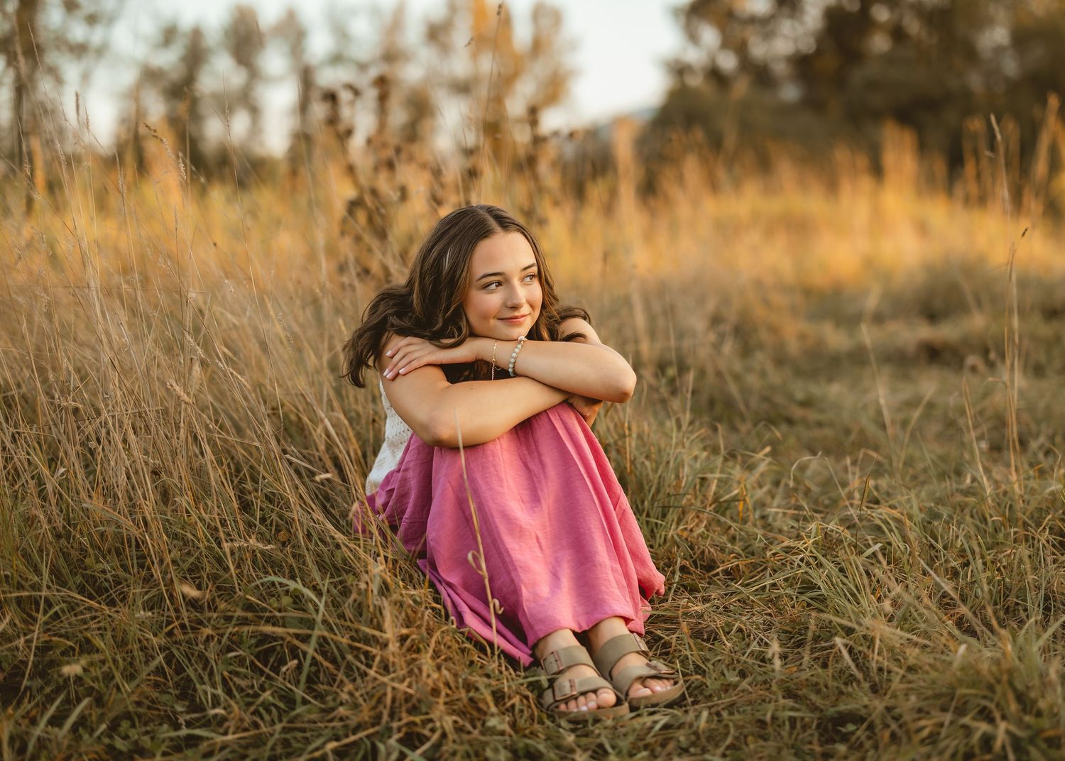 A person in a pink dress sits contentedly in a golden field of tall grass during sunset.