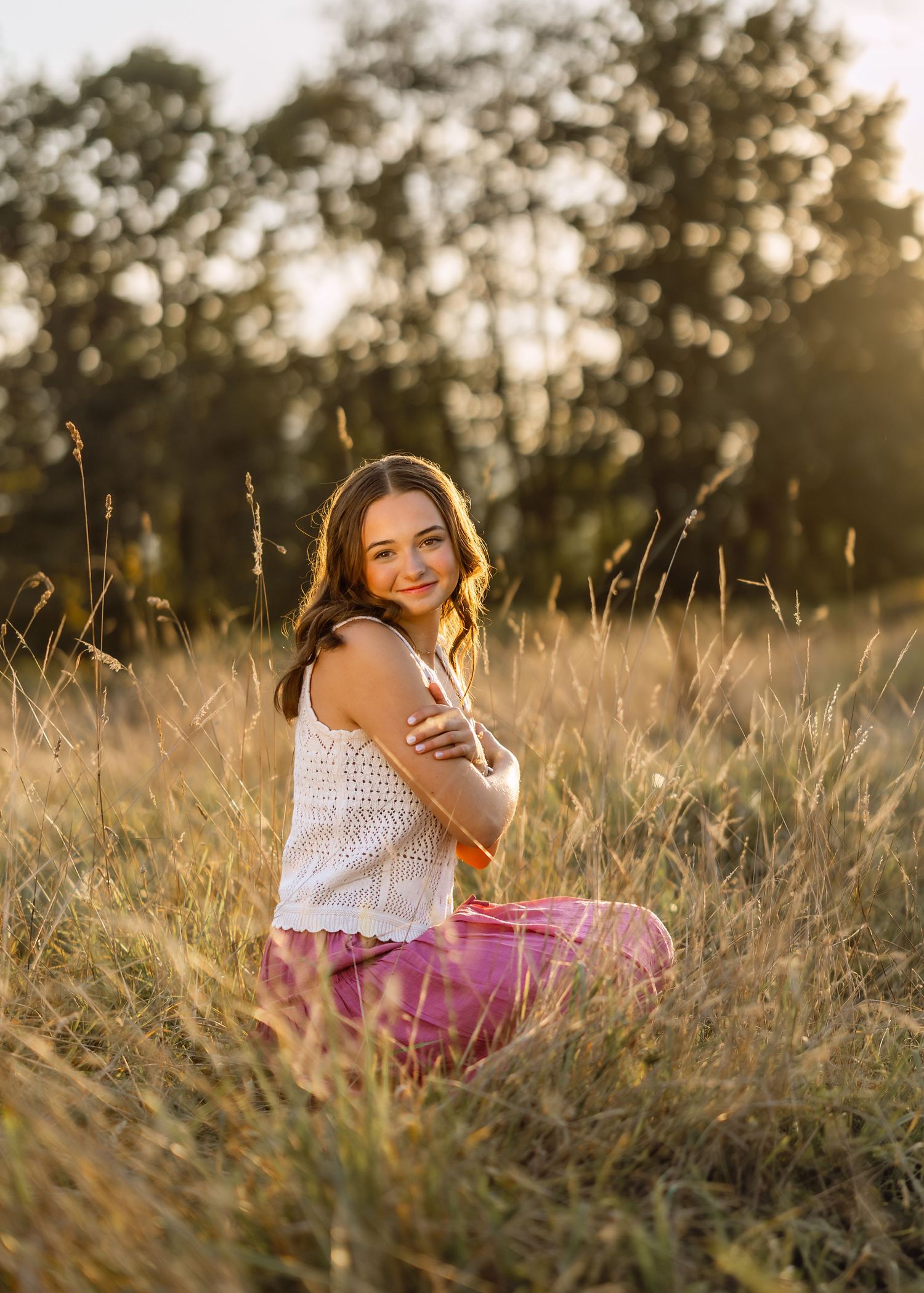 A person in a white top and pink skirt sits in a golden field of tall grass at sunset.