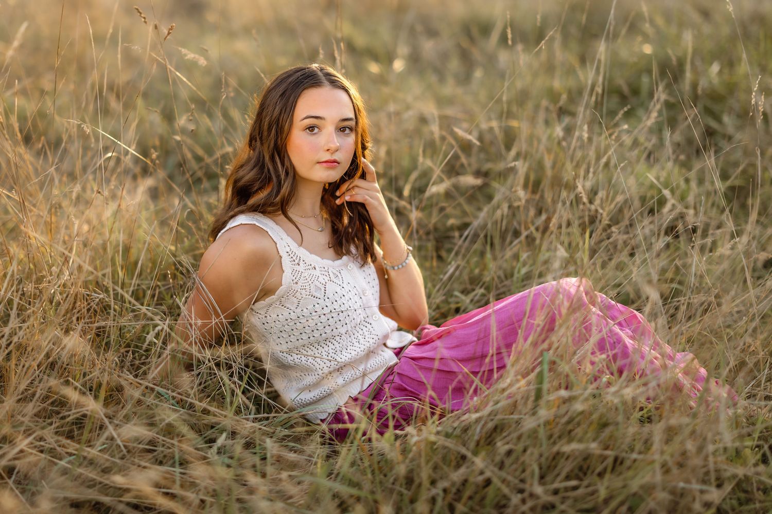 A person in a white tank top and pink skirt reclines in golden sunlit grass during a summer evening.