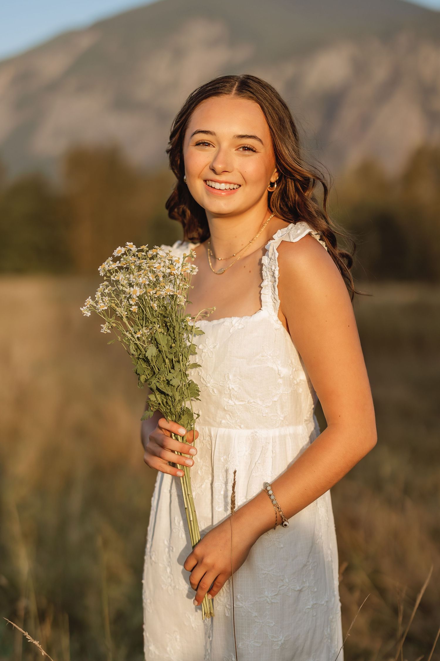 A person in a white sundress holds a bouquet of baby's breath flowers while posing in golden sunlight with mountains behind.