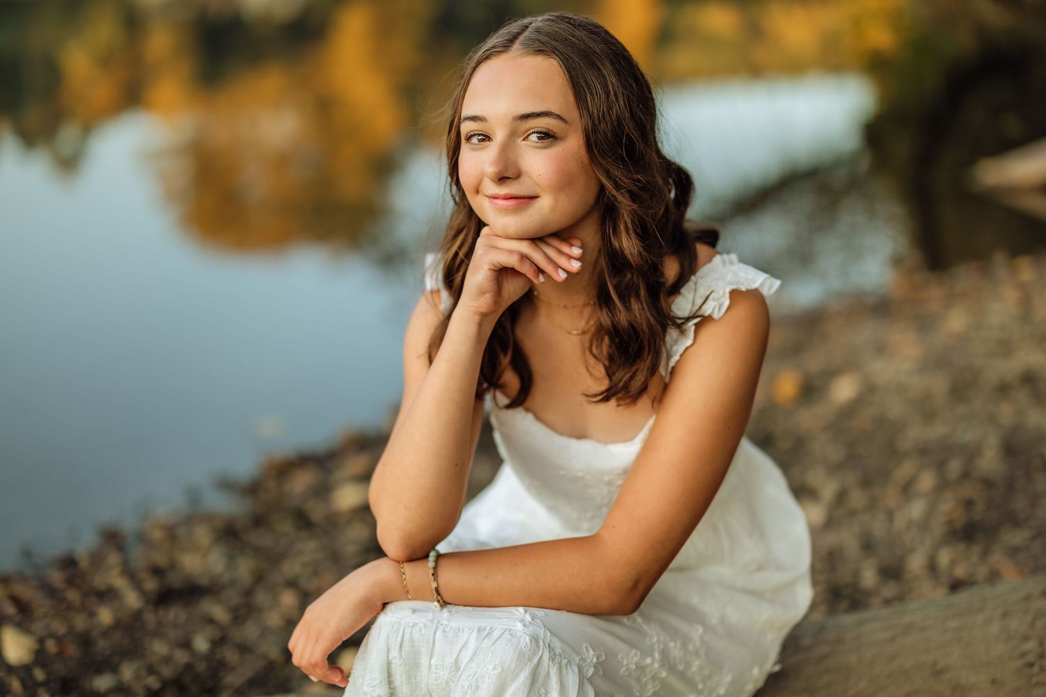 A person in a white dress sits thoughtfully by a lake during golden hour with autumn foliage in the background.