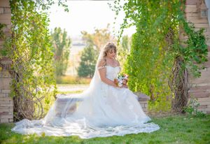 A bride in a flowing white dress poses in a garden setting with cascading vines and natural sunlight.