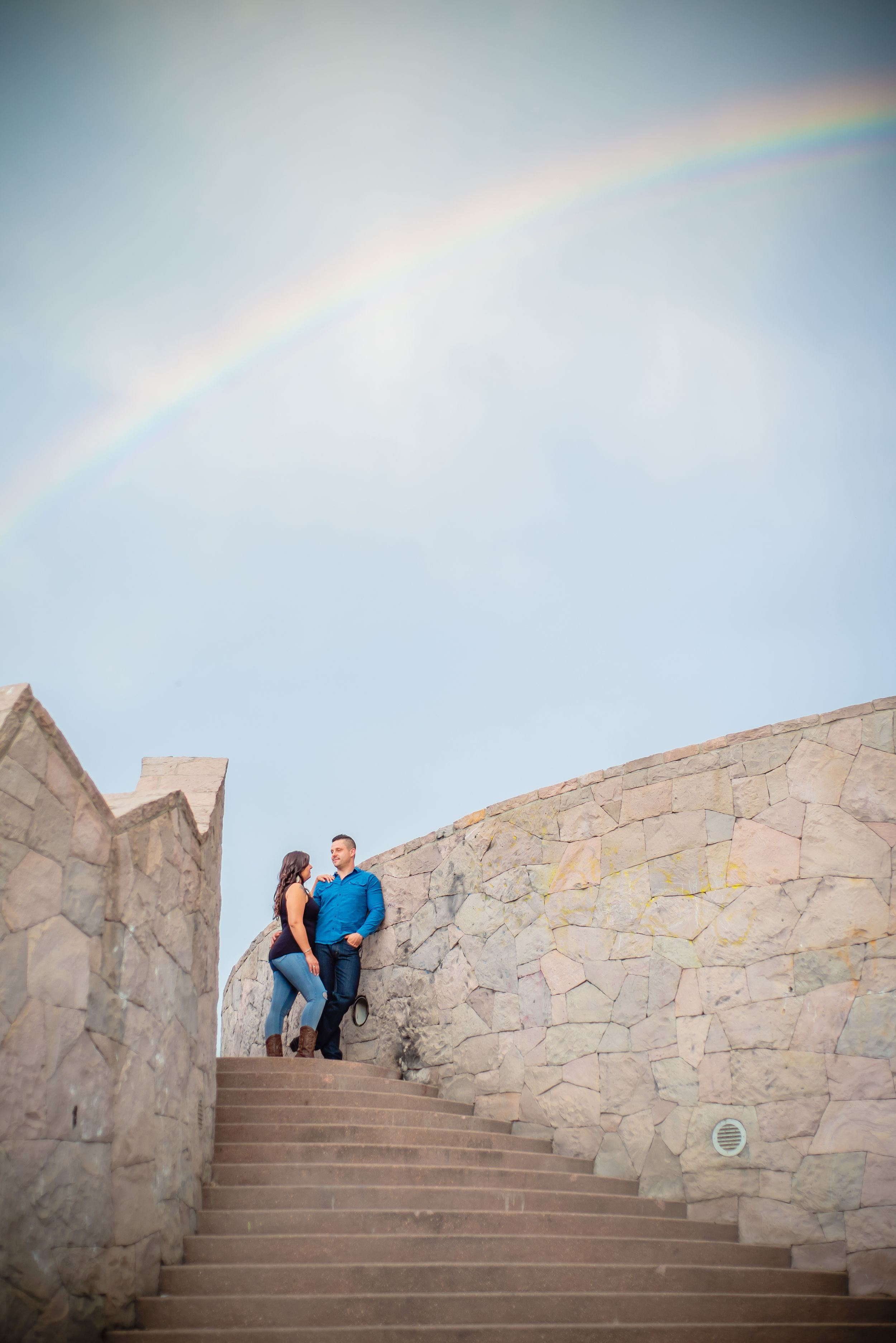 A couple stands on stone steps under a beautiful rainbow arch in the sky.