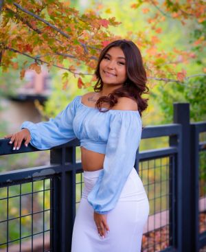 Portrait in blue crop top and white skirt against autumn foliage and metal railing.