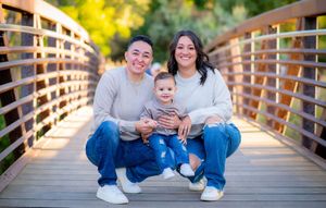 A family of three sits together smiling on a wooden bridge during sunset.