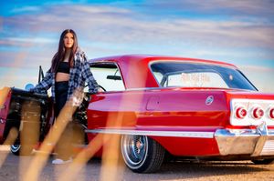 A vintage red Chevrolet Impala parked at sunset with rustic scenery in the background.