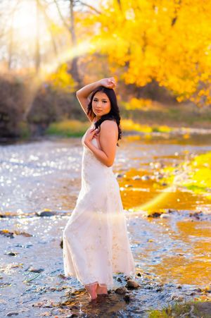 A white dress-clad figure poses by a stream with golden autumn foliage reflecting in the water.