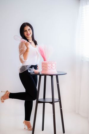 Baker poses with whisk next to pink cake on black table in white studio setting.