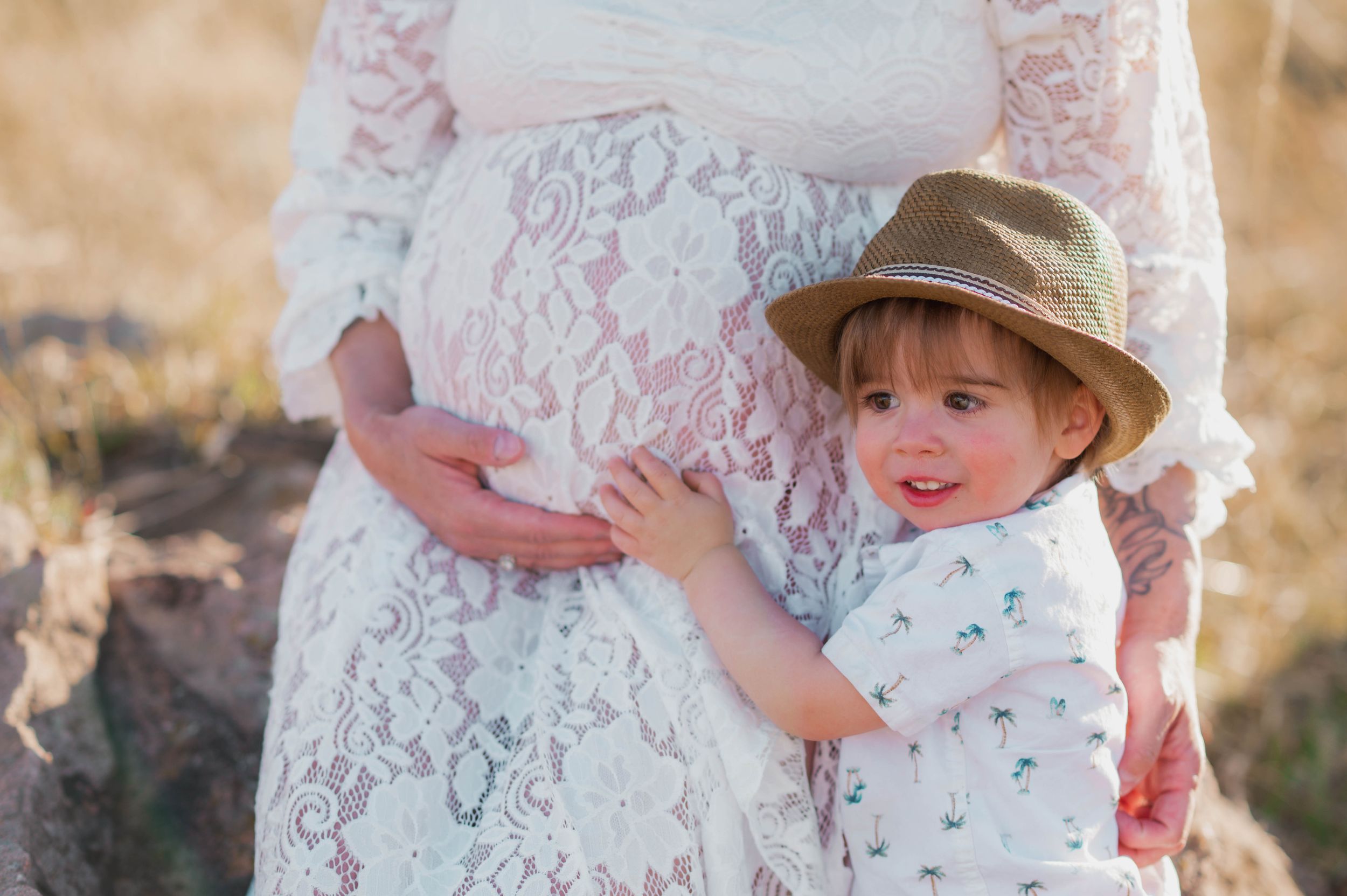 A toddler in a hat hugs a pregnant woman's belly outdoors in a warm, sunlit setting.