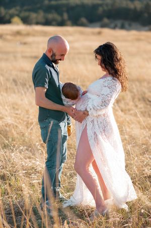 A couple stands in a golden field, lovingly cradling their newborn baby, the woman wearing an elegant white lace maternity gown.