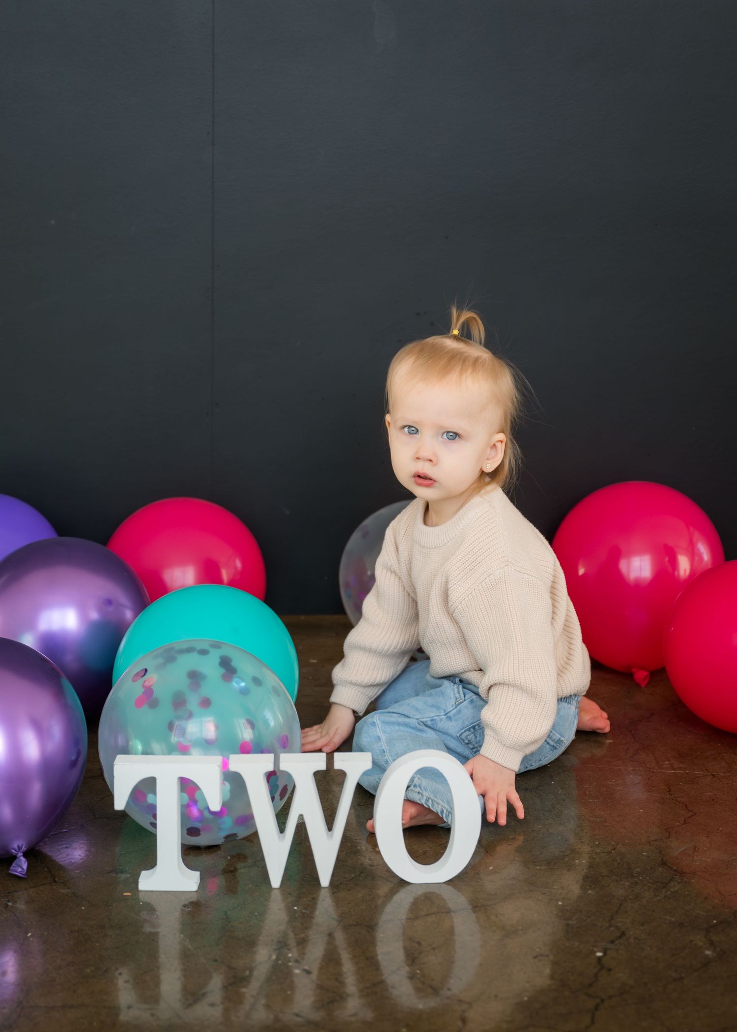 A toddler in beige sweater and jeans sits among colorful balloons next to large white 'TWO' letters against dark backdrop.