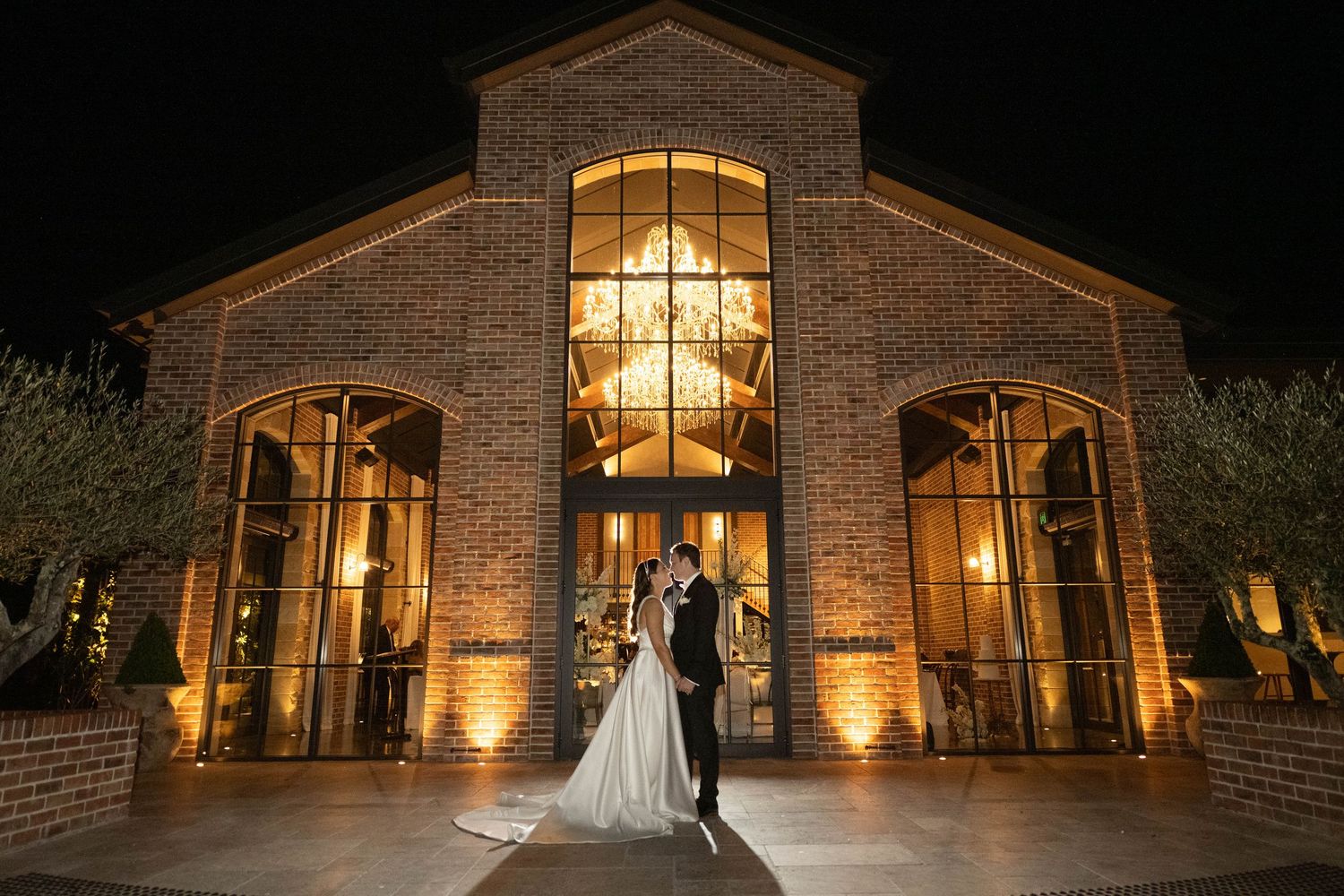 Bride and groom share romantic kiss outside illuminated brick wedding venue at night.