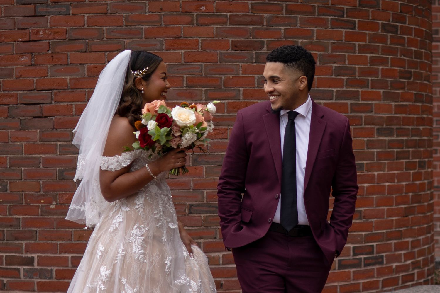 A couple in wedding attire shares joyful moments against a brick wall backdrop during their special day.
