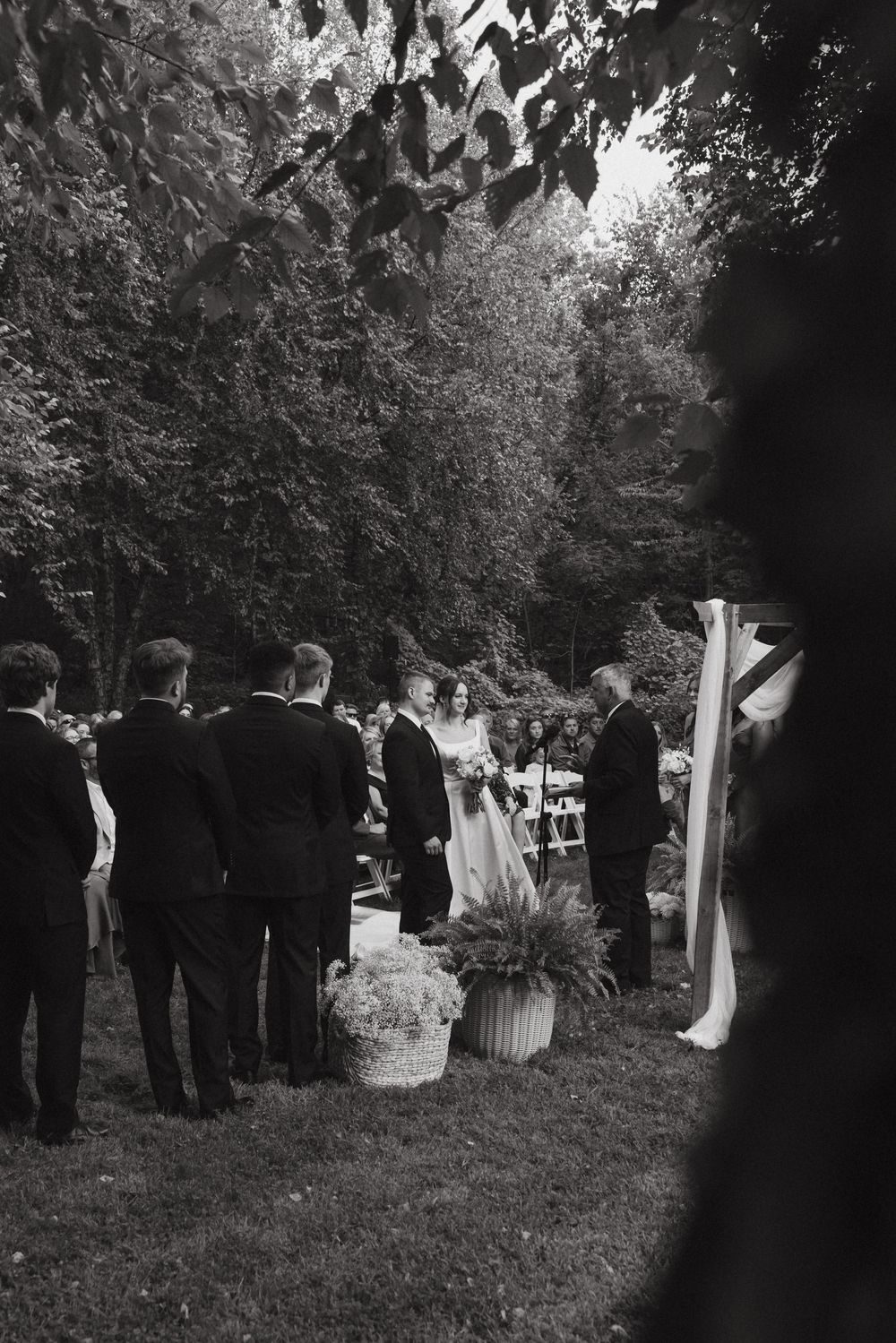 Black and white photo of an outdoor wedding ceremony with guests gathered under trees on a lawn.