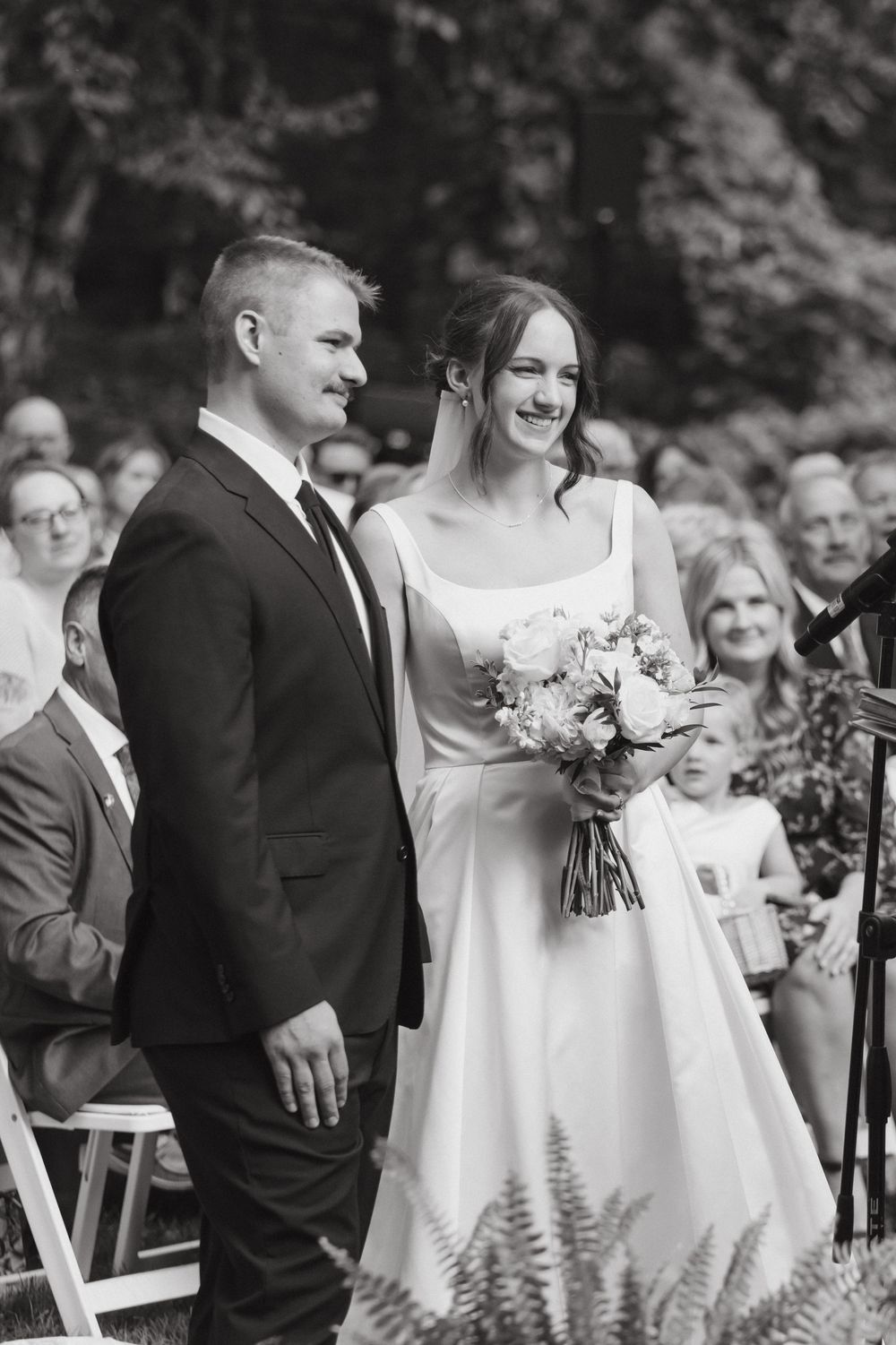A wedding ceremony in a wooded outdoor setting with guests seated on white chairs and floral arrangements.