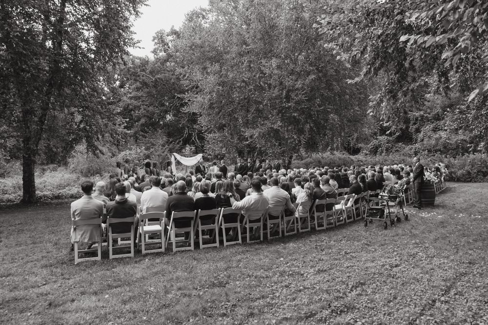 Outdoor wedding ceremony setup with wooden chairs arranged in rows beneath towering trees in a garden setting.