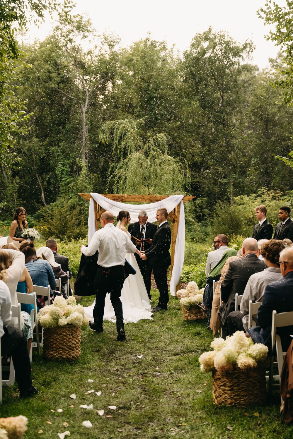 An outdoor wedding ceremony setup in a wooded area with white fabric draping and floral arrangements.