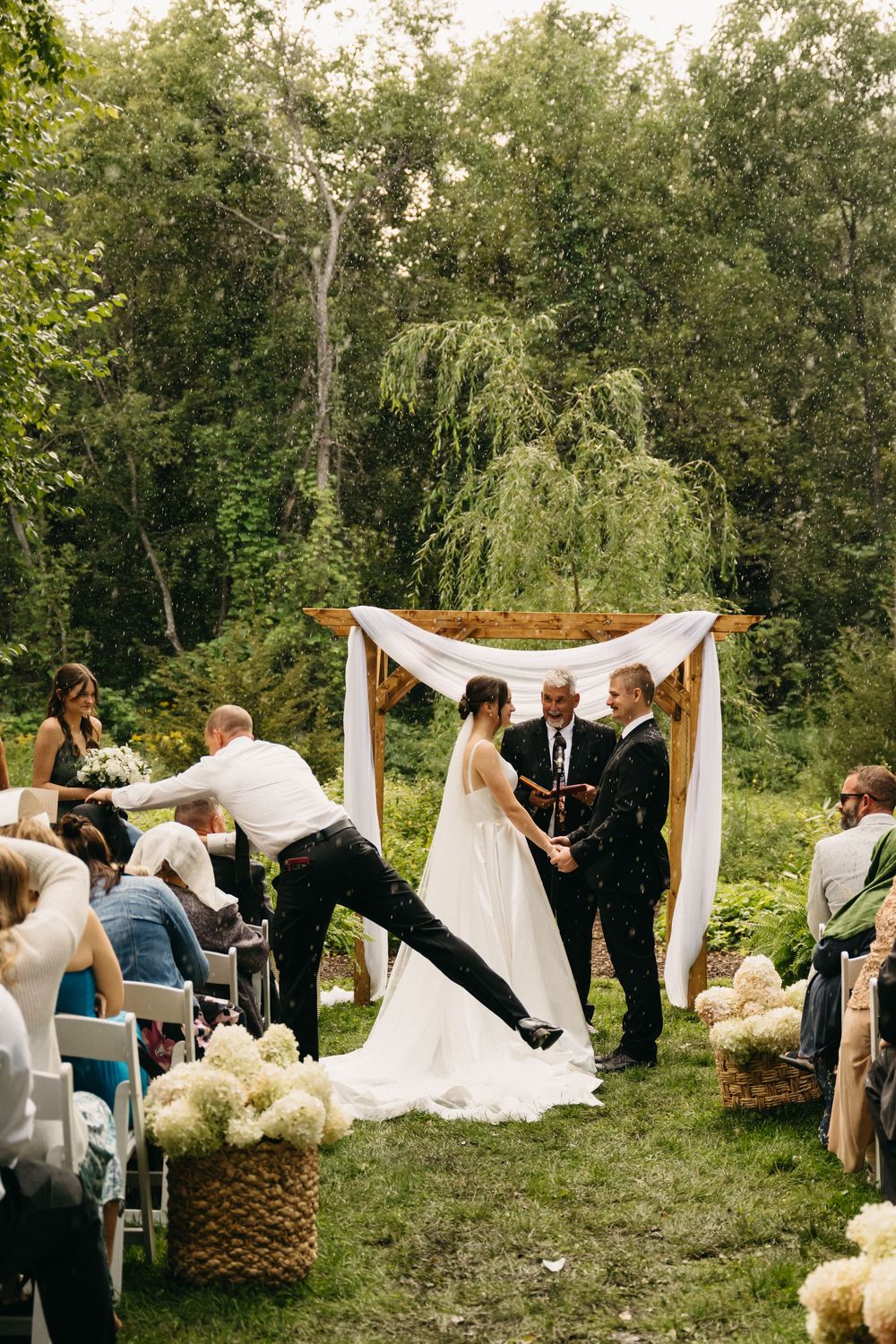 Outdoor wedding ceremony in a wooded setting with white fabric draped between posts and floral arrangements.