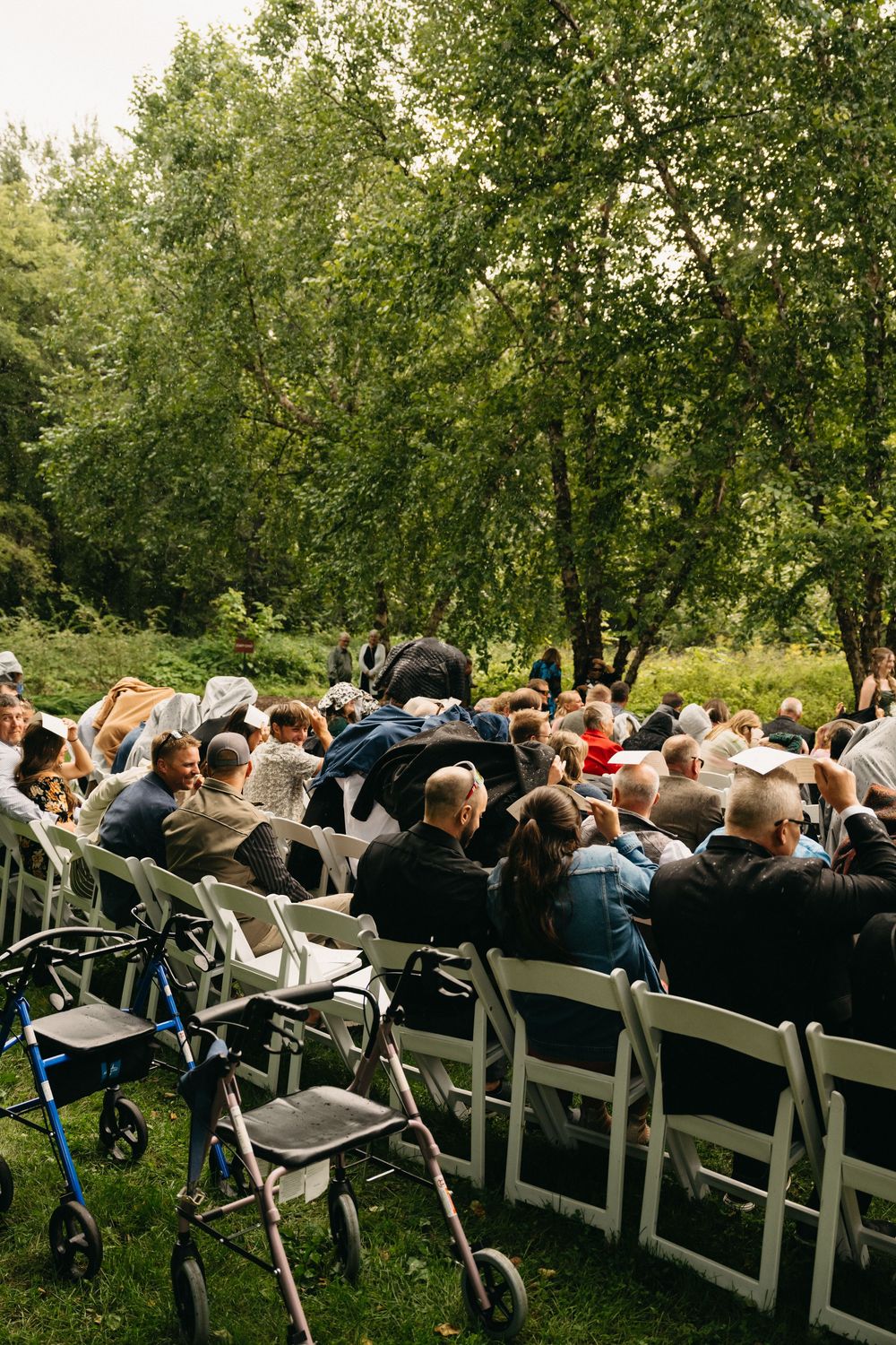 Guests seated in white folding chairs at an outdoor wedding ceremony surrounded by lush green trees.