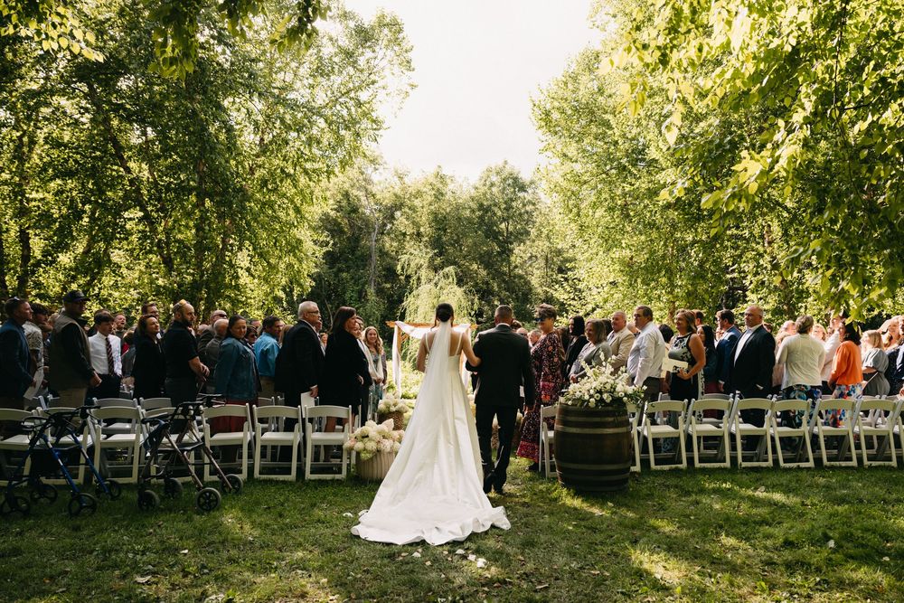 Outdoor wedding ceremony setup with white chairs arranged in rows under tall green trees on a sunny day.