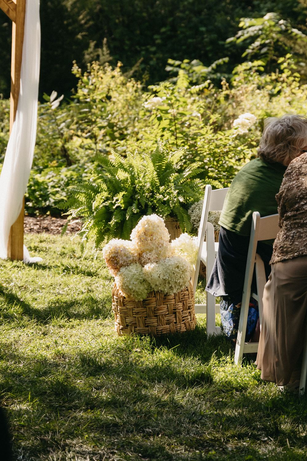 Rustic wedding ceremony decor features woven baskets with white hydrangeas lining an outdoor aisle in dappled sunlight.