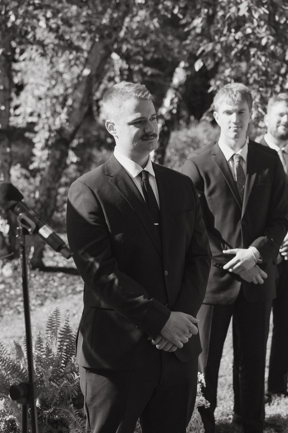 Group of men in black suits stand solemnly outdoors among trees in what appears to be a formal gathering.