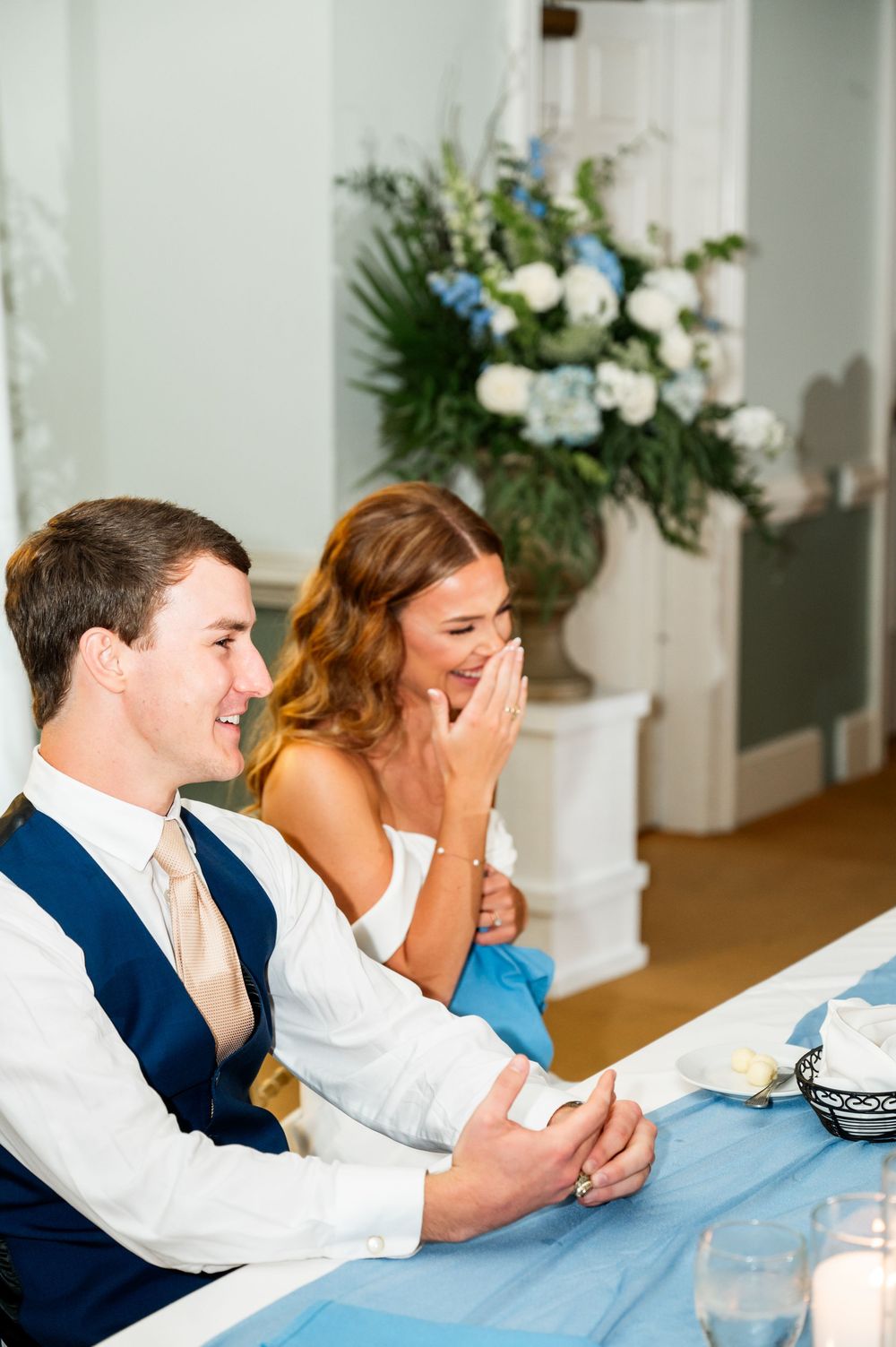 Wedding couple seated at head table during reception with floral decorations in background.