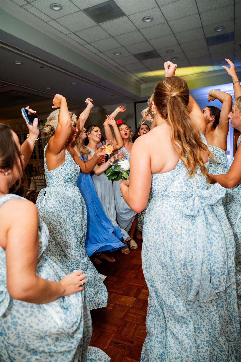 Bridesmaids in light blue floral dresses dancing together at wedding reception.