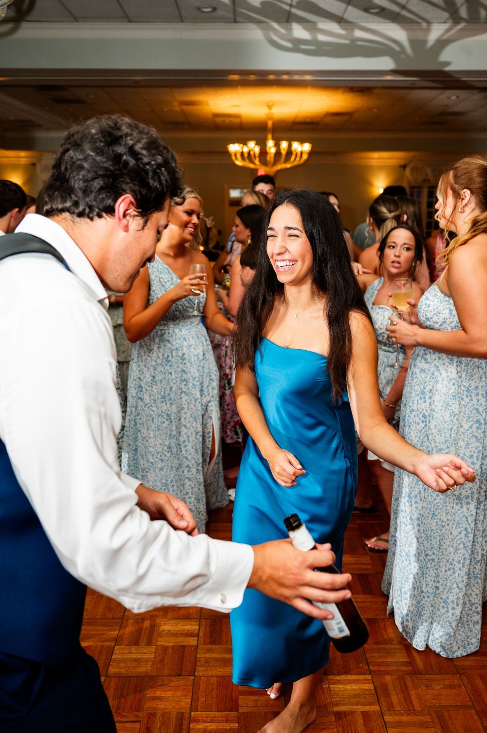 Wedding guests dancing together on parquet floor with chandelier lighting above.