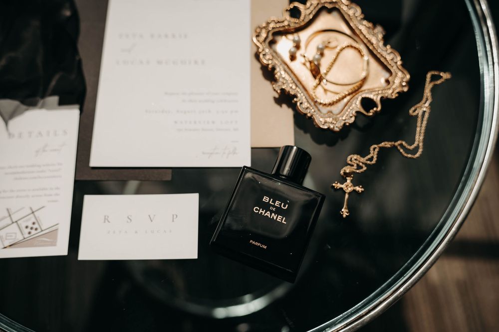 Bleu de Chanel perfume bottle beside gold jewelry tray and wedding invitation on glass table.