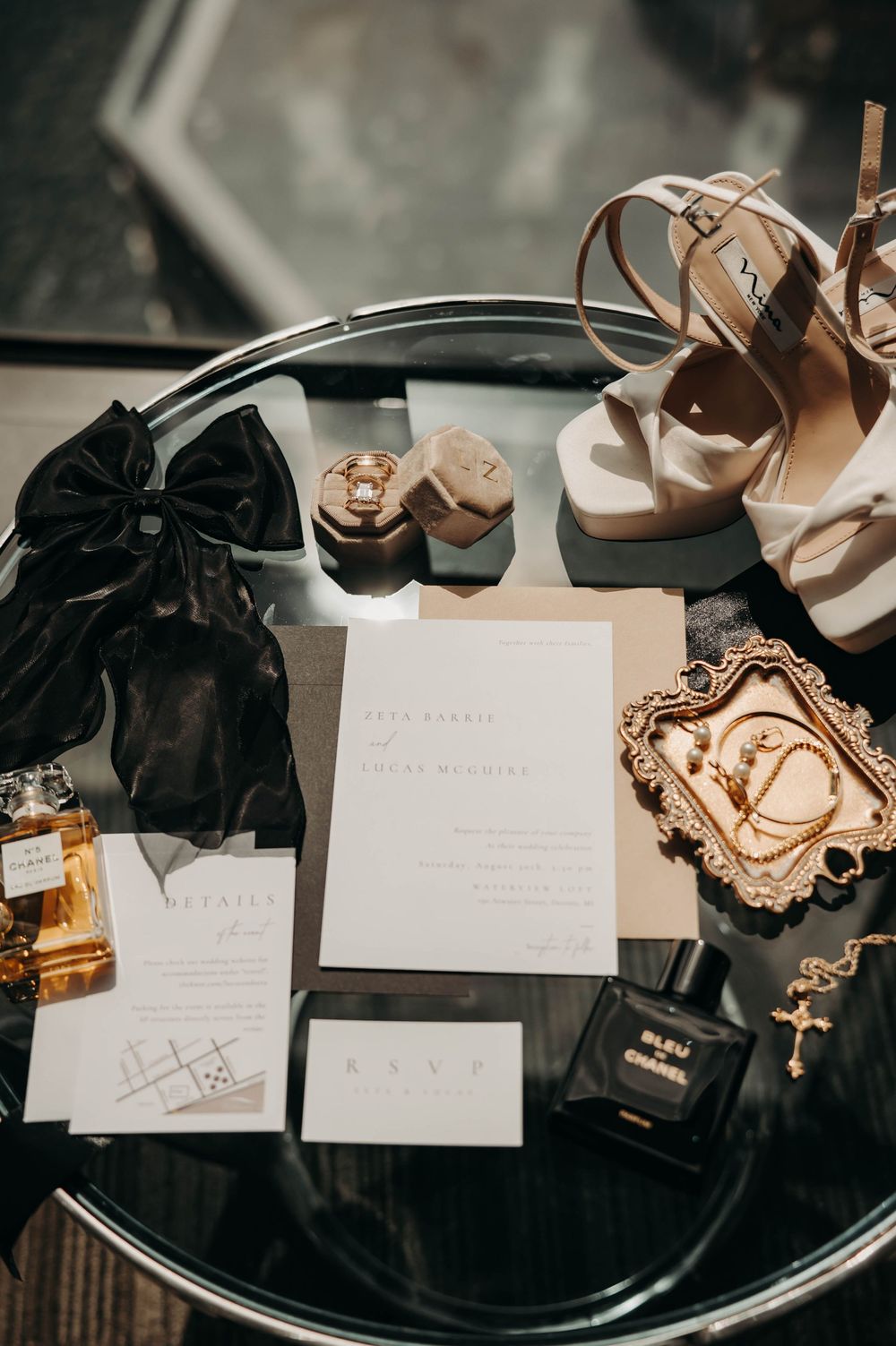 Elegant wedding flat lay with invitation suite, perfume, gold jewelry, heels, and black bow on glass table.