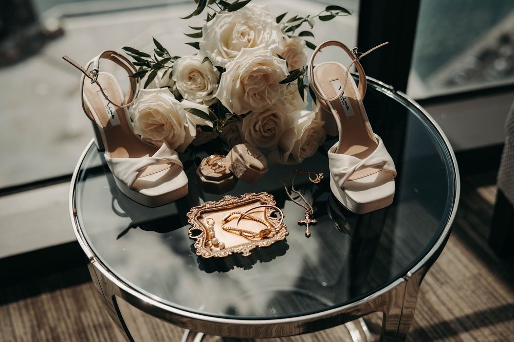Bridal flat lay featuring white heeled sandals, cream roses bouquet, gold rings, and cross necklace on glass table.