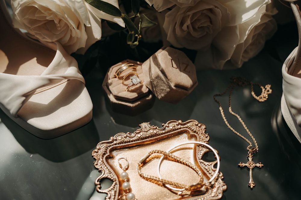 Close-up of gold wedding jewelry, diamond ring, cross necklace, and bridal heels with white roses on dark surface.